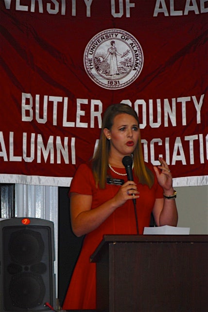 Lady in red dress standing behind podium with red Butler County Alumni Association banner in background.