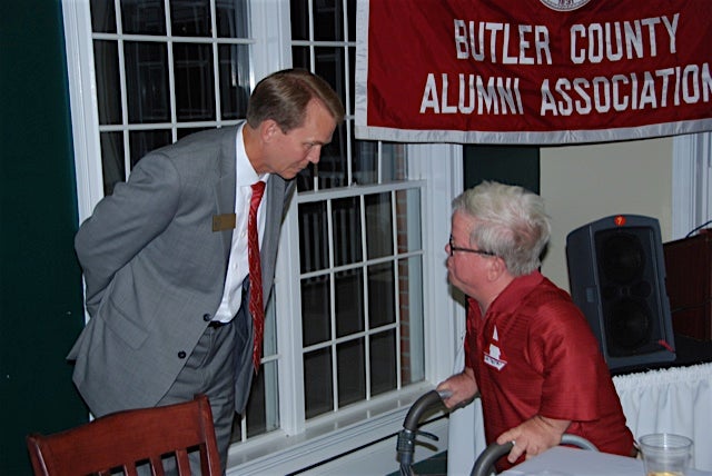 Man in gray suit and man in red shirt stanidng in front of window.