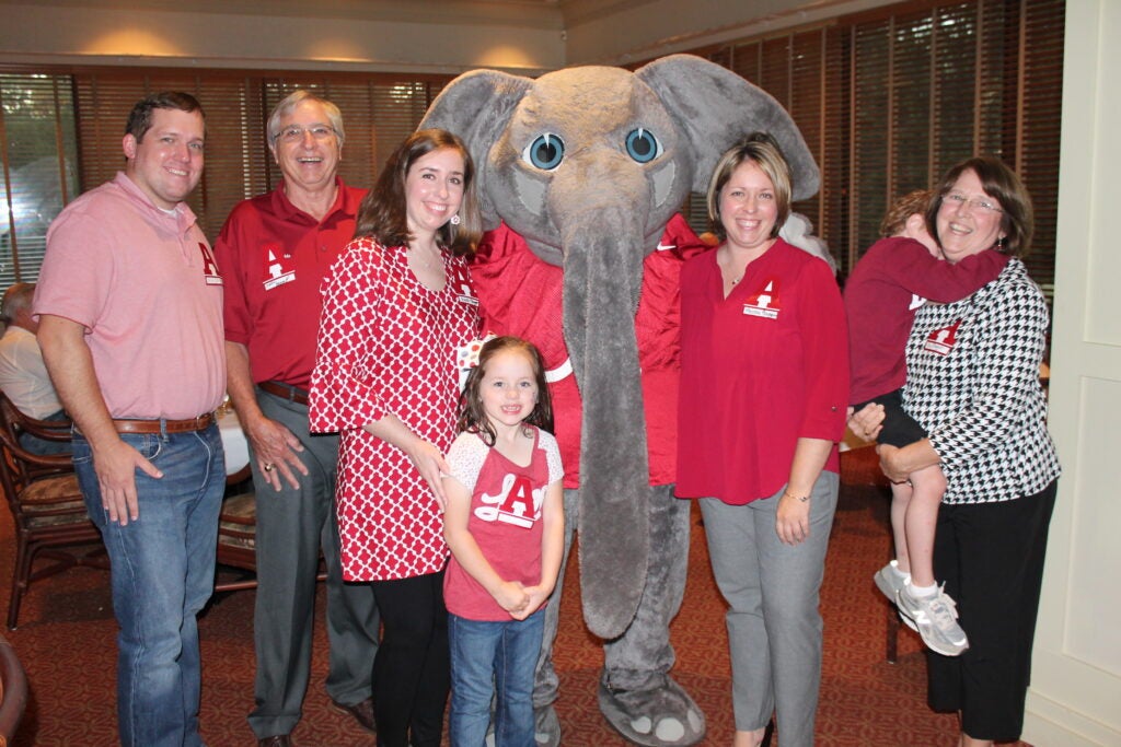 Group of people standing in room with Big Al Elephant in dim light room.
