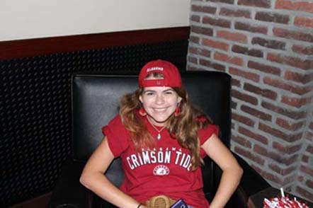 Girl sitting in brown chair with red shirt and cap on.