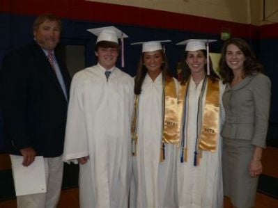 Group of students in white caps and gowns standing in front of dark wall.