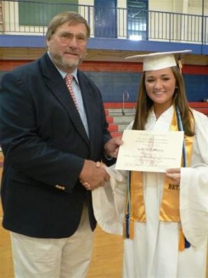 Man in blue jacket presenting certificate to student in white cap and gown.