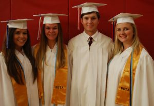Group of students with white cap and gowns on in front of red wall.