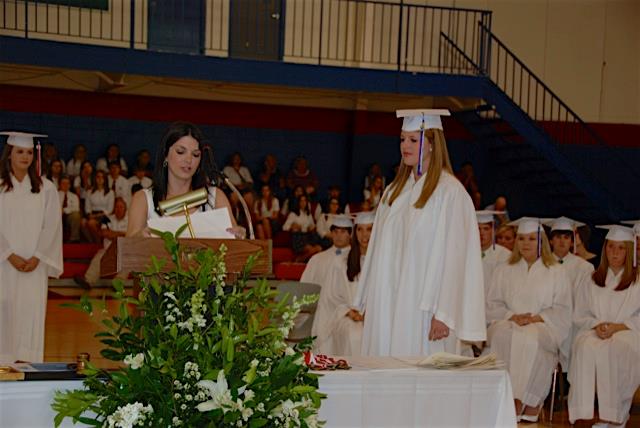 Lady standing behind podium in white top and students in the background in white caps and gowns.