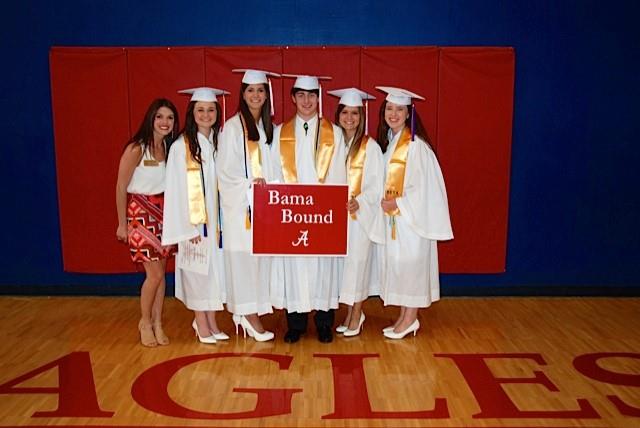 Group of students in white cap and gowns standing in front of red wall.