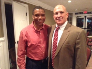 Man in red shirt and man in brown suit standing in a room.