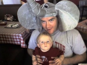 Man with Elephant Hat on holding a baby in crimson shirt.