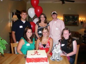 Group of people sitting at table with red and white cake.