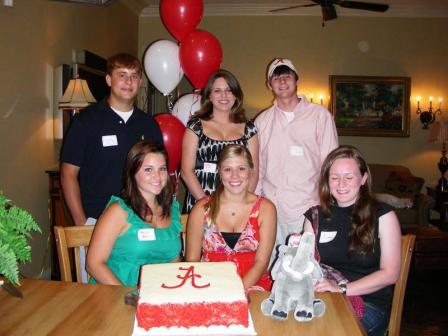 Group of people sitting at table with red and white cake.