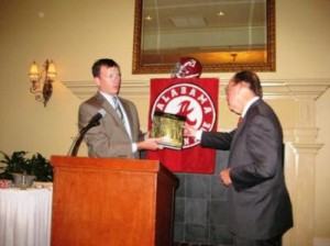 Man behind podium in gray jacket presenting an award to another person. Red banner in background.