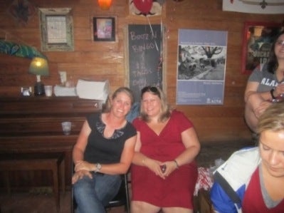 Two ladies sitting in room with brown wall. One lady standing in Blue/White jacket.