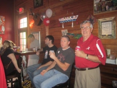 Two men sitting at at table and one man in red shirt standing.