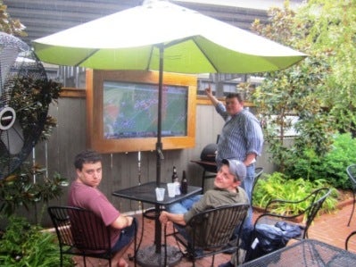Two men sitting at a patio table with umbrella and one man standing by a TV Screen outdoors.