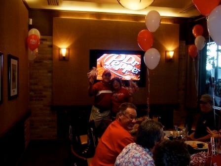 Group of people siting at tables in dim light room with TV screen in background.