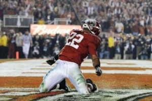 Alabama football player in red and white jersey kneeling on football field.