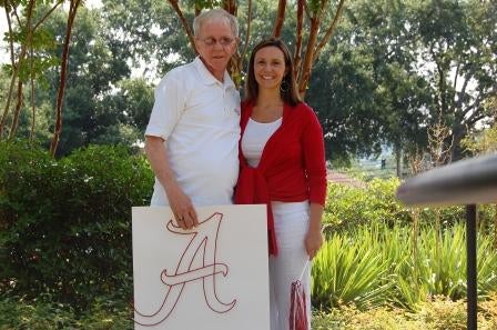 Man in white shirt and lady in red shirt standing outside holding a poster of an A.