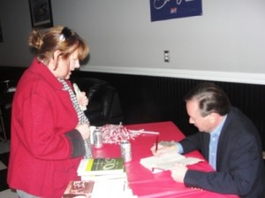 Women in red standing in front of table with red cloth. Man in Black jacket signing a book.