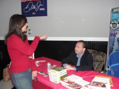 Women in red standing in front of table with red cloth. Man in Black jacket signing a book.