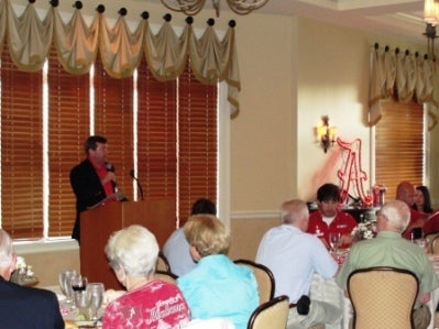 People in room sittting and Man in black jacket behind podium.