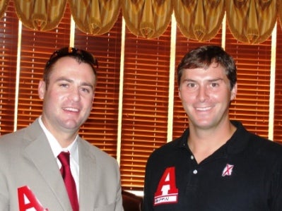 Two men standing in front of window with brown shades.