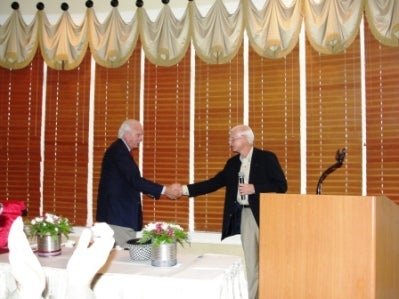 Two men shaking hands in front of table. Window with brown shades in the background.