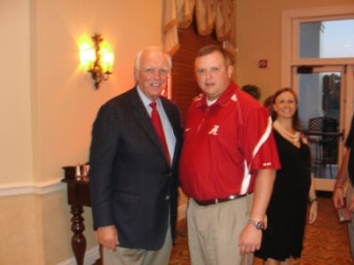 Man in black jacket and man in red shirt standing in room with dim light.