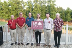 Group of people standing outside one in middle holding a red and white BAMA Bound sign.