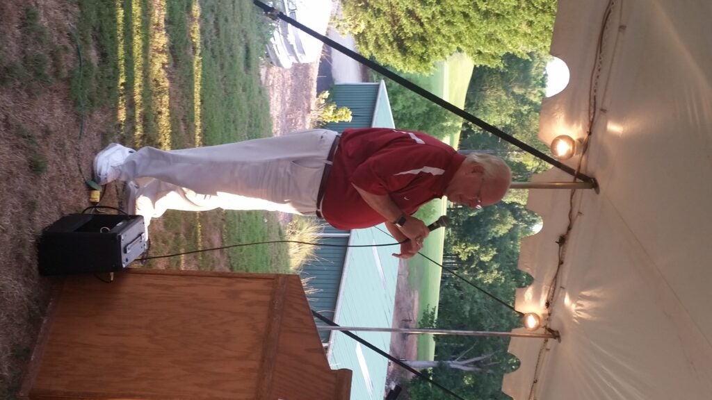 Man in red shirt and white pants standing outside holding a microphone.