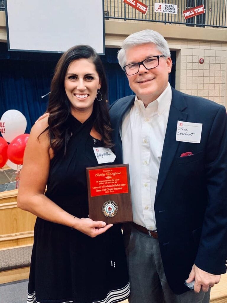 Lady in black dress holding an award and Man in black jacket standing in front of dark wall