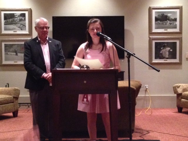 Man in dark suit and lady in pink dress standing behind podium in room with black/white wall.