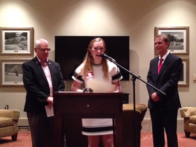 Two men in dark suits and lady in white dress standing behind podium with black/white wall.