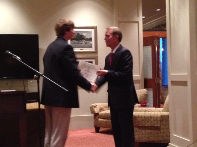 Two men in dark suits shaking hands in front of white wall.