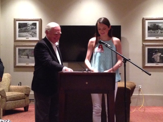 Man in dark suit and girl in green top standing behind podium with black/white wall.