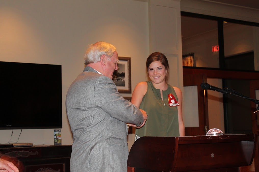 Man in gray jacket and lady in gray dress standing behind podium with white wall.