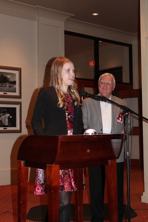 Lady in dark jacket standing at podium with man in gray jacket in dim light room.