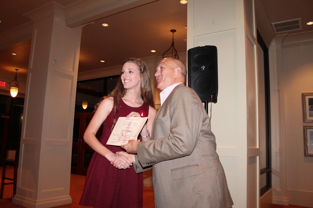 Dean Chuck Karr presenting a certificate to student in crimson dress in front of white/brown wall.