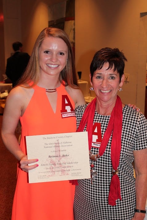 Student in orange dress holding a certificate and lady in black and white dress with red scarf.