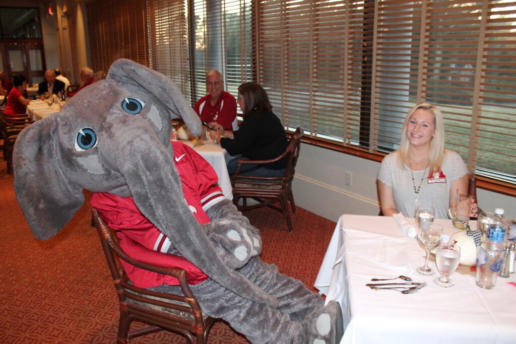Big Al Elephant sitting at table with lady in front of window with shades.