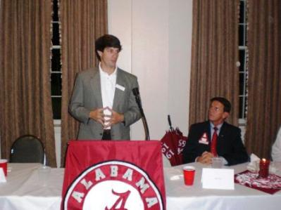 Man in Gray jacket standing behind podium speaking. Red banner on podium.