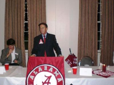 Man in black suit standing behind podium speaking. Red banner on podium.