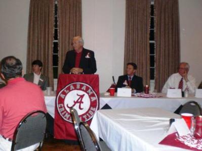 One person in black jacket behind podium speaking and goup of people sitting at table.