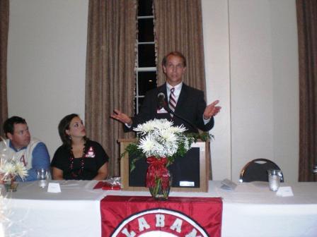 Man in black suit standing behind podium. People sitting at table beside the podium.