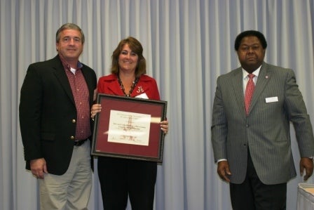 Group of people standing in front of gray curtain. One holding a framed picture.