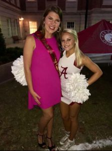 Two girls. One in pink dress and one in white Alabama outfit holding shakers.