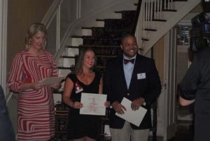 Two students holding certificates and lady in red and white dress standing in front of staircase.