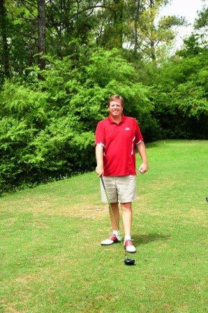 Man in red shirt standing outside with green trees in background.