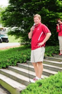 Man in red shirt holding a microphone standing on steps outside.