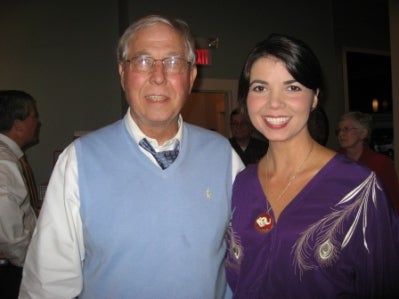Man in blue sweater vest and lady in purple top standing in dark room.