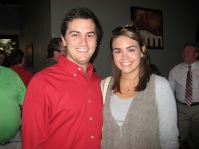 Man in red shirt and lady in beige top standing in dark room.