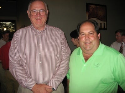 Man in beige shirt and man in green shirt standing in dark room.
